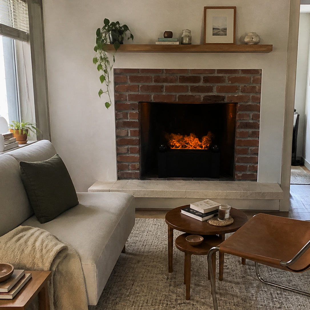 A cozy living room scene featuring a brick fireplace with a featuring a Aquafire Gatsby Water Vapor Fireplace, a light beige sofa with a dark green pillow, a brown leather accent chair, and a small wooden coffee table on a textured rug.